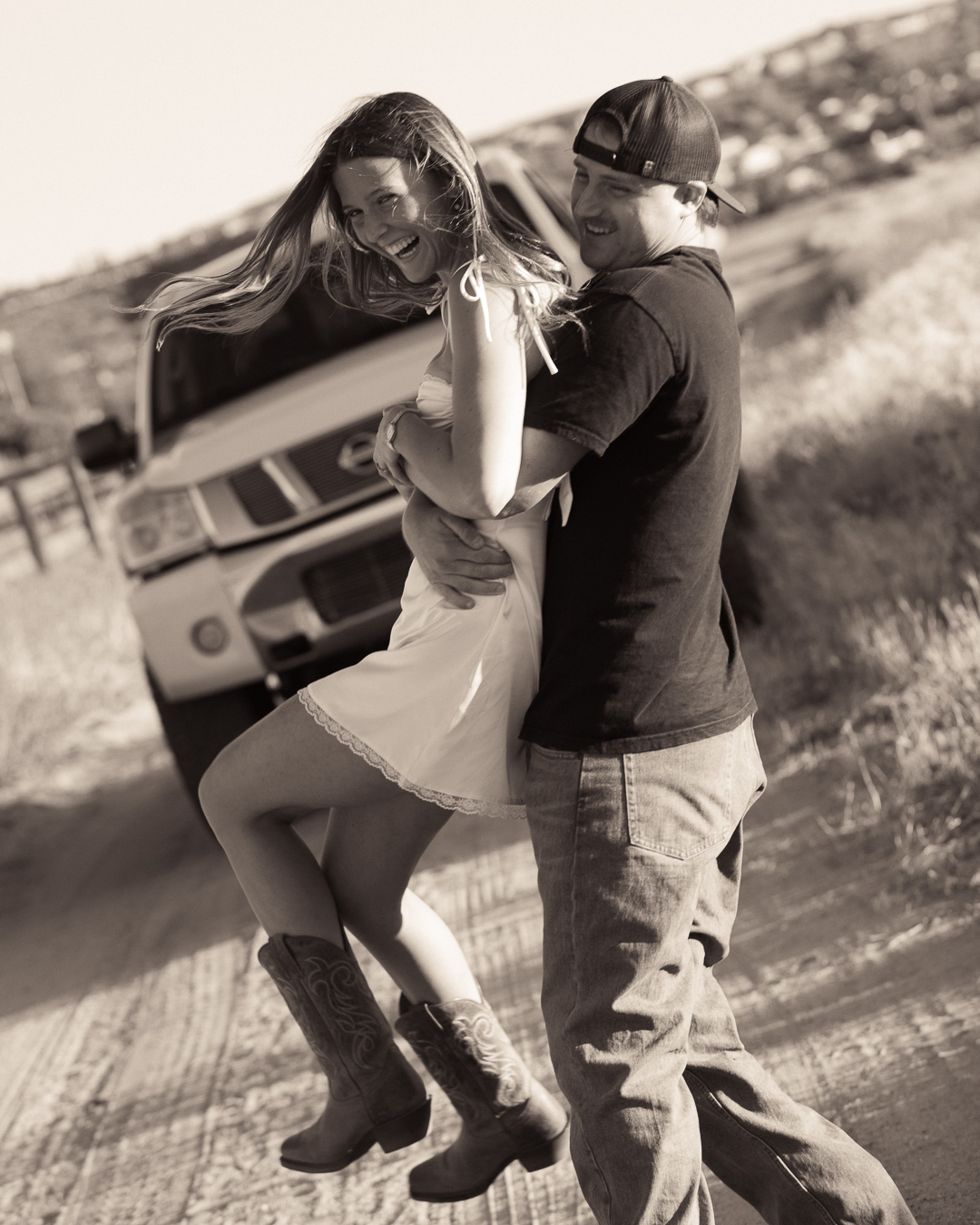 Black and white image of a couple laughing and dancing on a dirt road in front of a truck